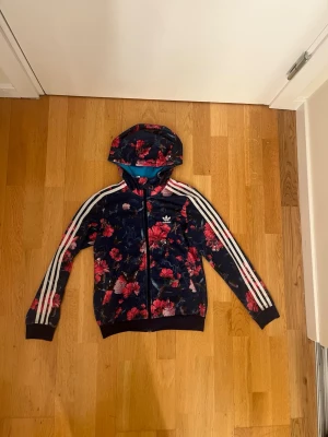 Adidas zip-hoodie med blommigt tryck - Adidas huvjacka med dragkedja och blommigt mönster för barn i storlek 152. Mörkblå med rosa blommor och tre vita ränder längs ärmarna. Har huva och fickor framtill.