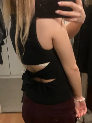 Svart ärmlös topp med knyt i ryggen - Säljer en svart ärmlös topp med snygg knytning och öppen ryggdetalj. Toppen har en croppad passform och är tillverkad i mjukt material som sitter skönt mot kroppen. Perfekt för dig som gillar stilrena plagg med en twist på ryggen.