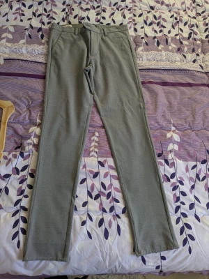 Grå kostymbyxor Jack & Jones Slim Marco - Slim Marco kostymbyxor från Jack & Jones i grått med smal passform. Klassisk design med bälteshällor, sidfickor och knappstängning. Materialet är mjukt och har en lätt stretch för extra komfort. Perfekt för en clean och stilren look.