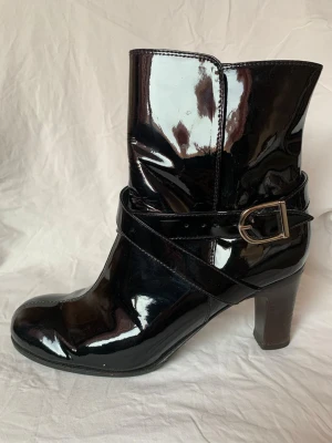 Svarta lackstövletter med klack - Säljer ett par svarta stövletter i glansigt lackmaterial (tror allt är läder) med rund tå och hög klack. Skorna har snygga remmar med metallspänne runt ankeln som ger en cool detalj. Perfekta för dig som gillar statement boots med lite edge. Klackhöjd som högst 7,5 cm. Sparsamt använda men har lite skav på sulans kant samt klack, och lite veck i lacken som det ju blir. Helhetsintrycket är dock väldigt gott och mycket fräscht inuti. Lite små i storleken kanske. 