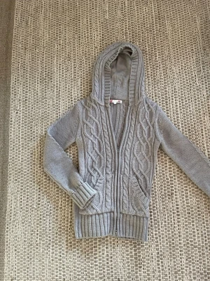 Grå stickad hoodie med dragkedja - Säljer en grå stickad hoodie med snyggt flätmönster både fram och bak. Tröjan har huva, långa ärmar och ribbade muddar vid ärmslut och nederkant.