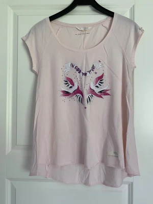 Ljusrosa t-shirt med fågeltryck från Odd Molly - Säljer en ljusrosa t-shirt från Odd Molly och en lång kofta från lager 157, t-shirt med coolt tryck av två vita och rosa fåglar och texten 'IN GOD WE TRUST' på bröstet. T-shirten har rund hals, korta ärmar och är tillverkad i mjuk bomull. Perfekt för dig som gillar unika prints och sköna material.