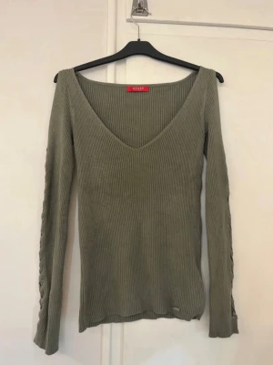 grön tröja - Snygg olivgrön ribbad tröja från Guess med v-ringad hals och långärmade ärmar. Tröjan har flätad detalj längs ärmarna som ger en cool touch. Perfekt att styla med jeans eller kjol för en trendig look.