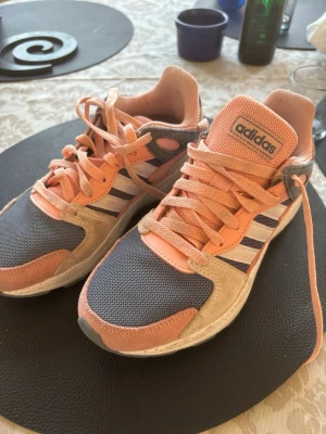 Adidas sneakers i persika och grå - Snygga Adidas sneakers med persikofärgade och grå meshpaneler, beige mockadetaljer och klassiska vita och grå ränder på sidan. Skorna har rund tå, platt sula och snörning. Perfekta för dig som gillar sportig stil och vill sticka ut med färg.