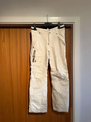 Vita Gore-Tex  från Peak Performance - Säljer ett par vita täckbyxor från Peak Performance i storlek M. Byxorna har flera praktiska fickor med dragkedja, markerade sömmar och en loose passform. Perfekta för vintern och skidåkning. Snygg design med detaljer i svart och robust material.