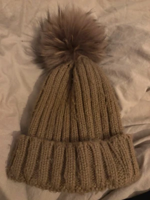 Beige stickad mössa med pälsboll - Säljer en beige stickad mössa med bred ribbad kant och en stor fluffig pälsboll på toppen. Perfekt för kalla dagar och ger en mysig vibe. Mössan är mjuk och har en klassisk design som passar till de flesta outfits. Tofsen går ta av. 
