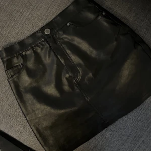 Svart skinnimitation kjol från H&M - Säljer en svart kortkjol från H&M Divided i skinnimitation. Kjolen har klassisk femficksdesign, knapp och dragkedja framtill. Snygg och enkel att matcha med olika stilar. Perfekt för dig som gillar en edgy look.