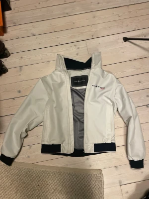 Vit vindjacka från Sail Racing - Snygg vit vindjacka från Salomon Racing med svart ribbad mudd vid ärmslut och nederkant. Jackan har dragkedja framtill, hög krage och diskreta loggor på bröstet och ryggen. Materialet är lätt och känns sportigt, perfekt för utomhusaktiviteter.