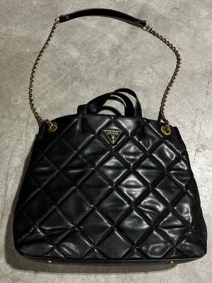 Svart quiltad handväska från Guess - Säljer en svart handväska från Guess med quiltad design och guldfärgade detaljer. Väskan har både kedjeaxelrem och handtag, samt flera fack med dragkedja inuti och utanpå, även ytterligare en mindre väska kopplad innanför. Materialet är skinn och insidan är fodrad med mönstrat tyg. Perfekt för dig som gillar stilrena accessoarer.