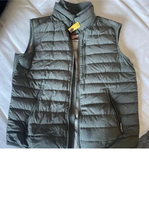 Parajumper väst - Säljer en grå dunväst från Parajumpers i storlek M. Västen är quiltad med horisontella sömmar, har ståkrage med gul detalj, två fickor med dragkedja och lättviktigt material. Perfekt för lager-på-lager och har en clean, sportig look.västen är äkta 