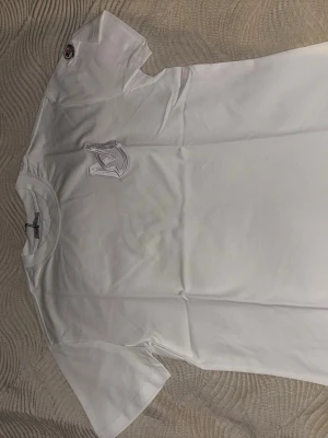 Vit t-shirt från Moncler med logga - Säljer en fräsch vit t-shirt från Moncler med broderad logga på bröstet och en patch på ärmen. T-shirten har rund hals och är kortärmad, tillverkad i mjuk bomull som känns skön mot huden. Perfekt för dig som gillar stilrena och exklusiva plagg.