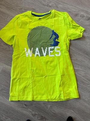 Gul t-shirt med surftryck från Pepperts - Cool gul t-shirt från Pepperts med blått och vitt surftryck och texten WAVES på bröstet. Rund hals och korta ärmar, perfekt för dig som gillar strandvibbar. Materialet är mjuk bomull som känns skönt mot huden.