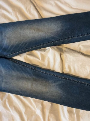 True Religion Geno Relaxed Slim jeans - Säljer ett par True Religion Geno Relaxed Slim jeans i klassisk blå tvätt med snygga slitningar. Jeansen har orange kontrastsömmar och den ikoniska hästskosymbolen på fickan. Modellen är slim men avslappnad och passar dig som gillar en modern look.