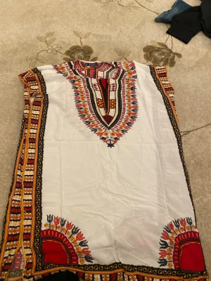 Vit mönstrad klänning från Lusa Paris - Säljer en vit klänning från Lusa Paris med färgstarka etno-inspirerade mönster i rött, svart, gult och orange. Klänningen har kort ärm, rund hals och detaljerade tryck längs kanterna och runt halsen. Perfekt för dig som gillar unika och färgglada plagg.