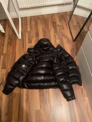 Moncler jacka  - Säljer en svart pufferjacka från Moncler med glansig finish och huva. Jackan har en klassisk Moncler-logga på ärmen, dragkedja framtill och ett coolt serietryck på insidan. Perfekt för dig som vill ha en varm och snygg jacka med streetkänsla. Den är även äkta och går att scanna. 