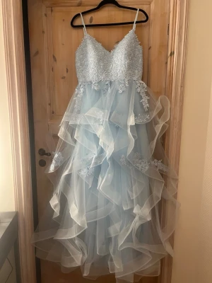 ljusblå Balklänning  - Superfin ljusblå långklänning med tunna axelband, spetsdetaljer upptill och flera lager volangig tyllkjol. Klänningen har v-ringning och blommiga broderier som ger en romantisk känsla. Perfekt för bal eller student! Materialet är mestadels tyll och spets.