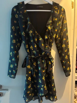 Mönstrad omlottklänning från Even&Odd - Snygg kort omlottklänning från Even&Odd med apelsiner i gult/orange på svart botten. Klänningen har volangdetaljer, v-ringning, knyt i midjan och långa lite genomskinliga ärmar. Jättesöt på. 