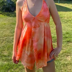 Orange omlott linnetopp med halterneck - Säljer en flowy omlott linnetopp i orange och ljusrosa toner. Toppen har djup v-ringning, tunna axelband som knyts i nacken och öppen rygg. Materialet är lätt och svalt, perfekt för varma dagar. Asymmetrisk nederkant ger en cool och unik look.