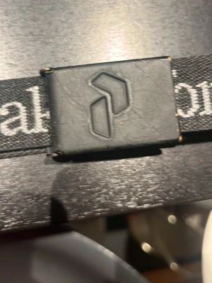 Peak Performance svart väst med logoband - Svart bälte från Peak Performance med ett tydligt logoband i svart och vitt. Västen har ett spänne framtill med Peak Performance-loggan och är gjord i ett slitstarkt syntetmaterial. Perfekt för dig som gillar sportig och clean stil. Kan vara lite svårt att stänga och öppna men är inga problem.