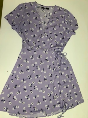 Ljuslila blommig omlottklänning Bik Bok - Supersöt ljuslila omlottklänning från Bik Bok i storlek XL. Klänningen har korta ärmar, knyt i midjan och ett vitt blommigt mönster över hela plagget. Tillverkad i mjuk polyester som känns skön mot huden. Perfekt för dig som gillar romantiska och feminina vibes.