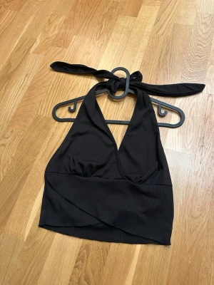 Svart topp - Säljer en svart omlott topp med knyt i nacken. Toppen är ribbad och har en djup v-ringning framtill. Materialet är mjukt och stretchigt, perfekt för att skapa en cool och trendig look. Passformen är croppad och djup i ryggen.