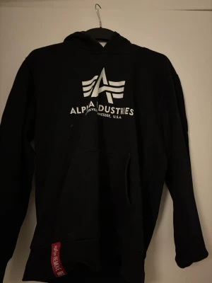 Svart hoodie från alpha industries - Det här är en klassisk svart hoodie från Alpha Industries med en stilren logotyp på bröstet. Den har en bekväm huva, en praktisk känguruficka och ribbstickade muddar. Priset kan diskuteras!