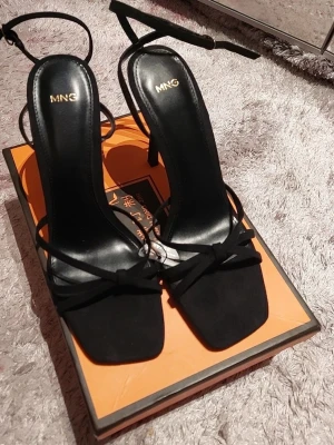 Svarta sandaler med klack från MNG - Snygga svarta sandaler från MNG med smala remmar och rosett framtill. Skorna har fyrkantig tå, hög klack och ankelsnörning. Materialet är en mix av skinn och mocka vilket ger en lyxig känsla. Perfekt för dig som gillar stilrena och trendiga skor.