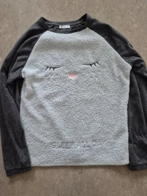 Tröja - Mysig grå fleece tröja från Cubus med lång ärm och fluffig framsida. Framsidan har ett broderat ansikte med stängda ögon och rosa näbb samt texten 'SLEEP ALL DAY' nertill. Perfekt för chill dagar hemma.