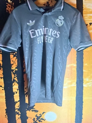 Grå Real Madrid adidas tröja - Säljer en grå Real Madrid tröja från adidas med klassisk krage och vita detaljer på ärmarna. Tröjan har broderat klubbmärke och adidas-logga på bröstet samt diskret mönster över hela tyget. Perfekt för dig som älskar fotboll och vill ha en snygg supportertröja.
