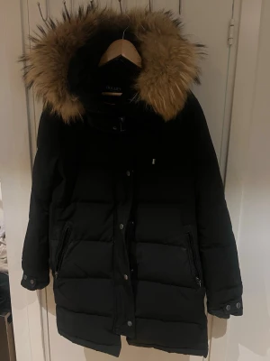 Svart pufferjacka med päls från Hollies - Svart pufferjacka från Hollies med stor fluffig päls på huvan. Jackan har dragkedja och knappar framtill, samt två fickor med dragkedja. Insidan har orange detaljer och jackan är perfekt för kalla dagar. Snygg och varm design som sticker ut.