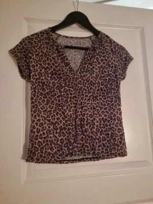Leopardmönstrad v-ringad topp - Säljer en kortärmad topp från H&M med leopardmönster i bruna och svarta toner. Toppen har v-ringad hals och är tillverkad i ett lätt och mjukt syntetmaterial. Perfekt för dig som gillar djurmönster och vill sticka ut lite extra.