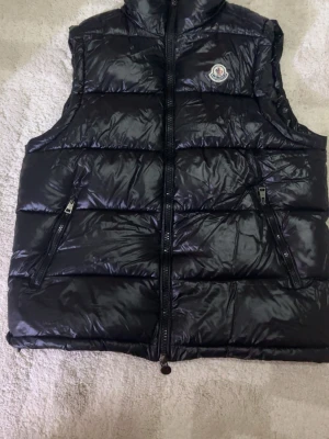 Svart dunväst från Moncler - Säljer en svart dunväst från Moncler med glansig finish och hög krage. Västen har två snedställda fickor med dragkedja och klassisk Moncler-logga på bröstet. Perfekt för lager-på-lager och riktigt snygg till höst- och vinterlooken.