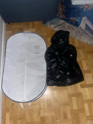 Svart Moncler pufferjacka med huva - Säljer en svart pufferjacka från Moncler med glansig finish och huva. Jackan har dragkedja framtill, två sidofickor med dragkedja och Moncler-logga på ärmen. Perfekt för dig som vill ha en snygg och varm jacka med exklusiv känsla. Inga skador på den köpte jackan för 1 vecka sen och säljer nu för att behöver pengar då kom gärna in med erbjudande 