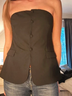 Festtopp  - Säljer en svart tubtopp med knappar framtill och snygga fickdetaljer. Kostymtoppen har en bandeauformad överdel och är figurnära med lätt utsvängd nederkant. Storlek M från H&M. Perfekt till nyår.