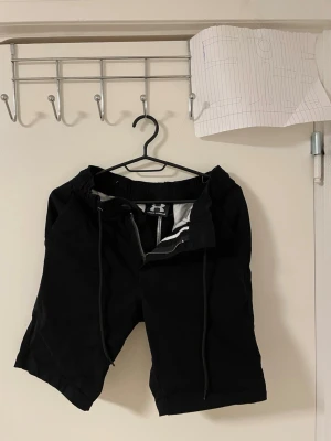 Svarta shorts Under Armour - Svarta shorts från Under Armour med snörning och dragkedja fram. De har elastisk midja och två bakfickor. Materialet känns som bomull och de har en clean, sportig look. Perfekta för chill eller träning. Liknande modell som originala.
