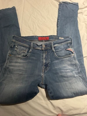 Replay Anbass ljusblå jeans, W30 L32 - Säljer ett par Replay Anbass jeans i ljusblå tvätt med snygga slitningar och klassiska fem fickor. Jeansen har normal passform och raka ben, med Replay-logga på fickan och läderpatch bak i midjan. Perfekt för dig som gillar en avslappnad stil.
