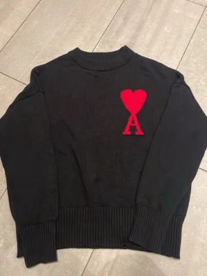Svart stickad tröja från Ami - Svart stickad tröja från Ami Alexandre Mattiussi med ribbad krage, muddar och nederkant. På bröstet finns ett stort rött hjärta och bokstaven A. Perfekt för dig som gillar stilrena detaljer med en twist.