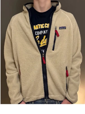 Patagonia fleeece - Säljer en beige fleecejacka från Patagonia med mörkblå detaljer och röda dragkedjor. Jackan har hög krage, tre fickor med dragkedja och en loose passform. Perfekt för dig som gillar chill och comfy stil.