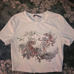Vit ribbad t-shirt med tigermotiv - Säljer en vit ribbad t-shirt med korta ärmar och ett coolt tryck av två tigrar bland blommor och växter på framsidan. T-shirten har en rund halsringning och är croppad i modellen, perfekt för dig som gillar unika prints.