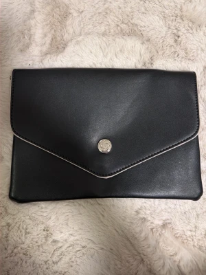 Svart kuvertväska i skinnimitation - Stilren svart kuvertväska i skinnimitation med silverfärgad knapp framtill. Väskan har ett enkelt fack inuti och stängs med tryckknapp. Perfekt till att bära det viktigaste och matcha med olika outfits.