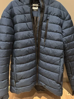 Blå pufferjacka från McKinley - Säljer en blå skidjacka från McKinley i storlek medium. Jackan har huva, flera dragkedjefickor och quiltad design med horisontella sömmar.
