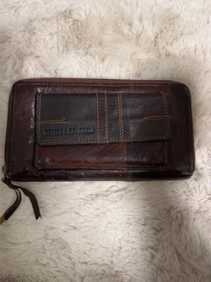 Brun plånbok från Spikes & Sparrow - Snygg brun plånbok i skinn från Spikes & Sparrow med flera fack och dragkedja runtom. Framsidan har en dekorativ ficka med kontrastsömmar och logotyp. Insidan har gott om plats för kort, mynt och sedlar. Perfekt för dig som gillar klassisk stil.