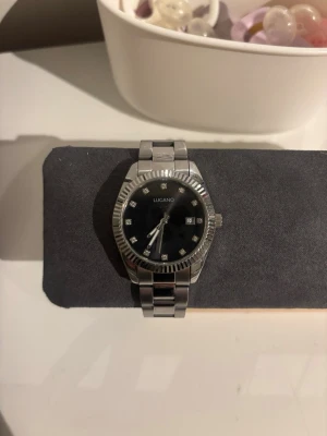 Klocka  - Säljer en klocka från Lugano i rostfritt stål med svart urtavla och zirkoner. Fullt fungerande och i fint skick då den endast blivit använd 1 gång. Info om klockan finns på sista bilden 
