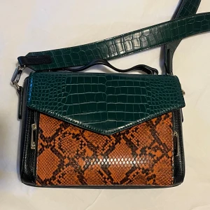 BECKSÖNDERGAARD Grön och brun axelväska  - Säljer en unik axelväska från Becksöndergaard  med grönt krokodilpräglat lock och axelrem, samt brun ormskinnsmönstrad framsida. Väskan har guldfärgade metaldetaljer och flera fack. Perfekt för dig som gillar statement-accessoarer och coola mönster.