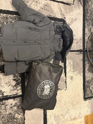 Mörkgrå pufferjacka från Canada Goose - Säljer en mörkgrå pufferjacka från Canada Goose med snyggt broderat märke på ärmen. Jackan har huva, tryckknappar och är fodrad med dun för extra värme. Perfekt för kalla dagar och har en clean, modern look. Medföljer originalpåse och tag.