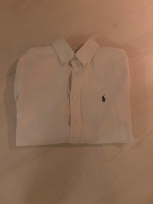 Vit skjorta från Ralph Lauren - Säljer en klassisk vit skjorta från Ralph Lauren med ikonisk broderad logga på bröstet. Skjortan har knappar hela vägen och en snygg krage med knappdetaljer. Storlek 16 som ska mot svara en xs/xxs.