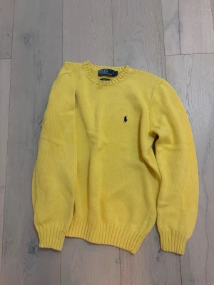 Gul stickad tröja från Polo Ralph Lauren - Säljer en gul stickad tröja från Polo Ralph Lauren med klassisk rund hals och ribbade muddar. Tröjan har en broderad logga på bröstet och är långärmad. Perfekt för dig som vill ha en färgglad och stilren look.