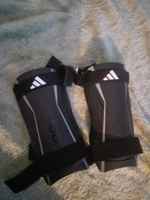 Adidas benskydd svart med vita detaljer - Säljer ett par svarta Adidas benskydd med vita detaljer och justerbara kardborreband. De har en sportig design med Adidas-loggan på framsidan och är gjorda i slitstark syntet. Perfekt för dig som spelar fotboll och vill ha bra skydd på planen.