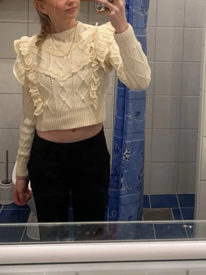 Beige stickad croppad tröja med volang - Superfin beige stickad tröja med volangdetaljer och spets längs axlar och bröst. Croppad modell med lång ärm och ribbade muddar. Perfekt för dig som gillar romantisk och trendig stil.