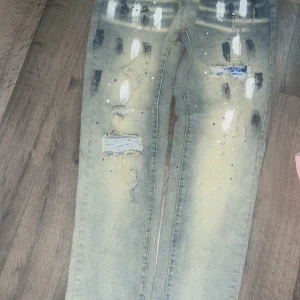 Blå jeans från Purple Brand med färgstänk - Säljer ett par blå jeans från Purple Brand med coola färgstänk i vitt och svart samt slitna detaljer och hål. Jeansen har klassisk femficksdesign, knappstängning och en ljus tvätt med unika effekter. Perfekt för dig som gillar streetstyle och vill sticka ut.